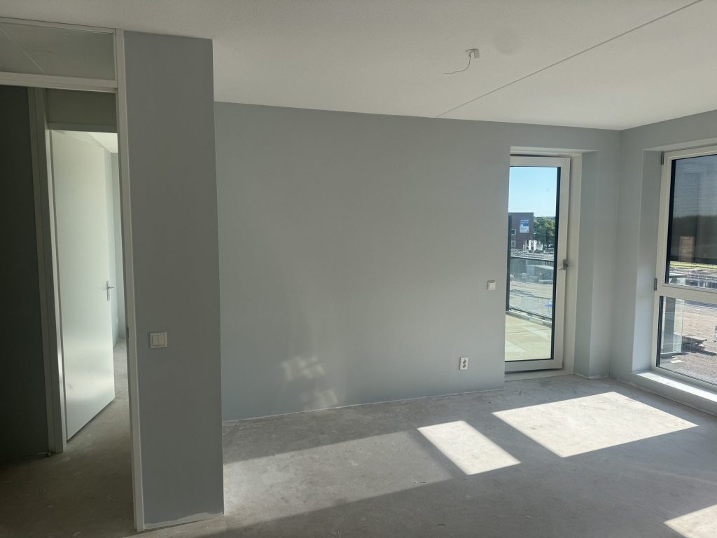 Nieuw appartement met kale, strakke wanden, een betonvloer en een balkondeur met zicht op een bouwterrein