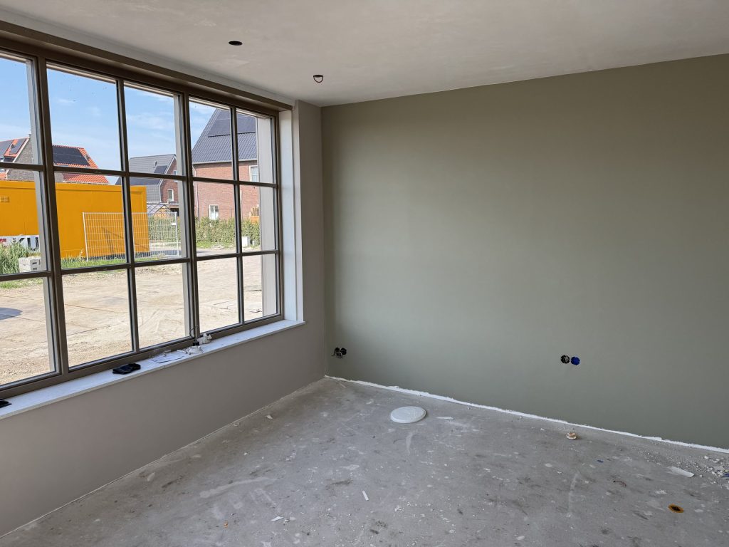 Kamer in een nieuw huis met een egaal afgewerkte grijsgroene wand en een groot raam met zicht op nieuwe woningen