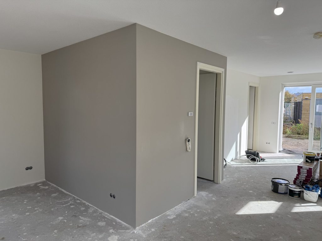 Woonruimte in een nieuwbouwwoning met een pas gesausde grijze wand, een betonvloer en verfemmers bij de tuindeur
