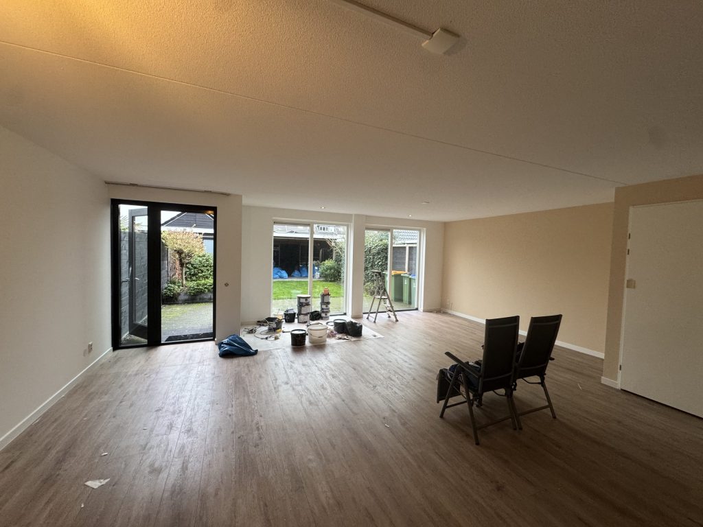 Ruime woonkamer van een nieuwe eengezinswoning met lichte wanden, een houten vloer, tuindeuren en verfemmers op de vloer