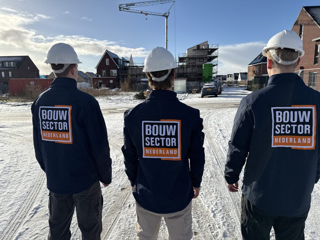 Drie vakmensen van Bouwsector Nederland met witte helmen kijken naar nieuwbouwwoningen in aanbouw met een bouwkraan erboven